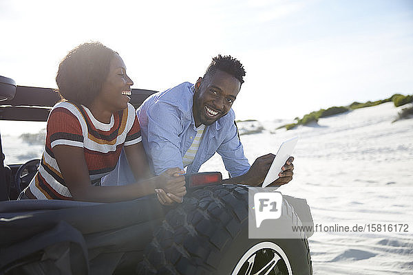 Lächelndes junges Paar benutzt digitales Tablet im Jeep am sonnigen Strand