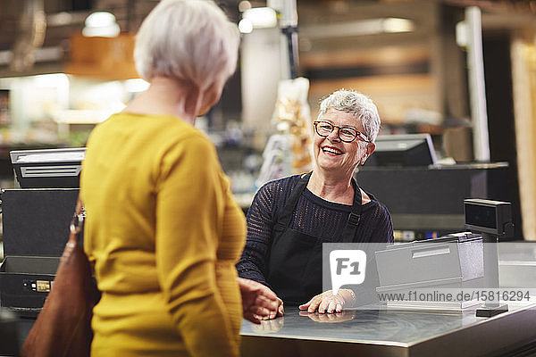 Happy senior female cashier greeting customer at supermarket checkout