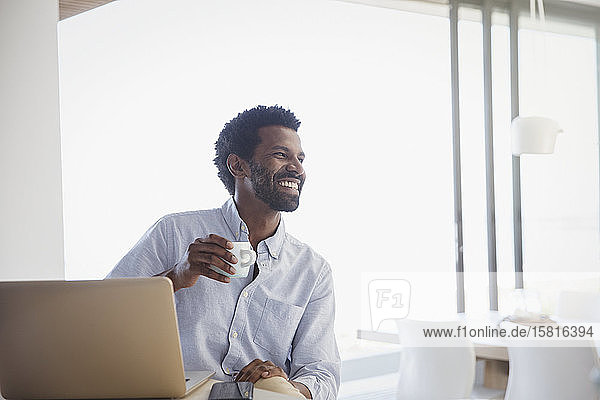 Lächelnder  begeisterter Mann  der Kaffee trinkt und zu Hause am Laptop arbeitet