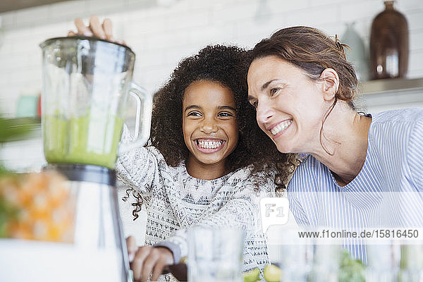 Lächelnde  begeisterte Mutter und Tochter bei der Zubereitung eines gesunden grünen Smoothies im Mixer in der Küche