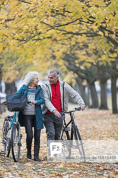 Smiling senior couple walking bicycles among trees and leaves in autumn park