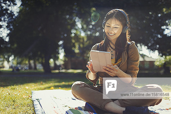 Lächelnde junge Frau  die ein digitales Tablet benutzt und mit Kopfhörern im sonnigen Sommerpark Musik hört