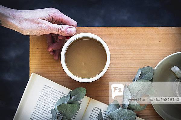 Frauenhand hält eine Kaffeetasse auf einem Holztisch mit einem Buch