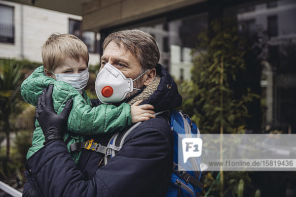 Vater trägt Sohn, beide tragen Schutzmasken, im Freien