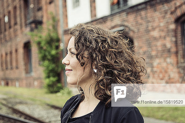 Lächelnde brünette Frau mit lockigem Haar vor einem Backsteingebäude und alten Eisenbahnschienen