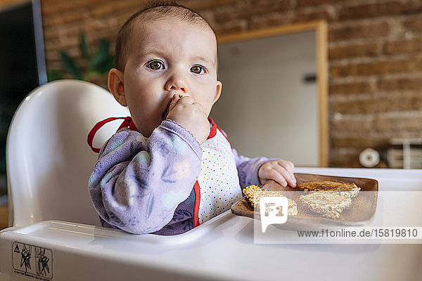 Porträt eines kleinen Mädchens auf einem Hochstuhl sitzend  das hausgemachte Haferflockenplätzchen mit den Händen isst