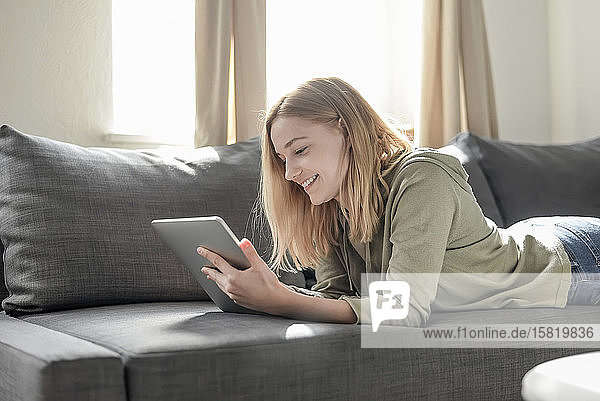 Porträt einer lächelnden jungen Frau  die zu Hause auf der Couch liegt und ein digitales Tablet benutzt
