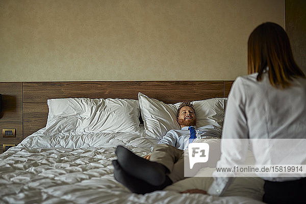 Businessman lying on bed in hotel room looking at businesswoman