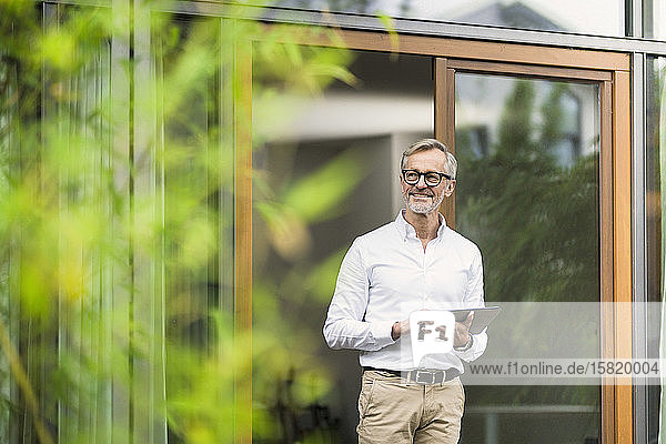 Lächelnder älterer Mann mit grauen Haaren hält Tablette vor seinem modernen Design-Haus im Bambusgarten