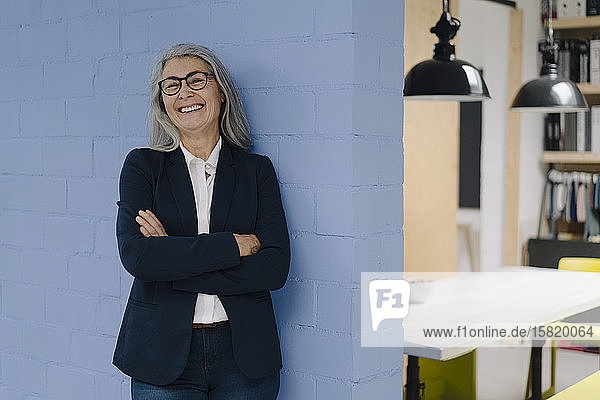 Porträt einer glücklichen grauhaarigen Geschäftsfrau  die an einer blauen Wand in einem Loft-Büro steht
