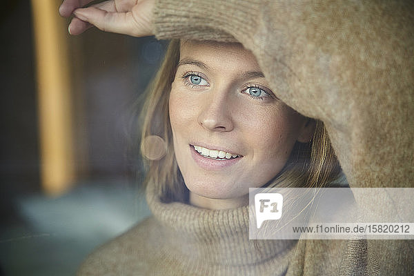 Porträt einer lächelnden blonden jungen Frau hinter einer Fensterscheibe