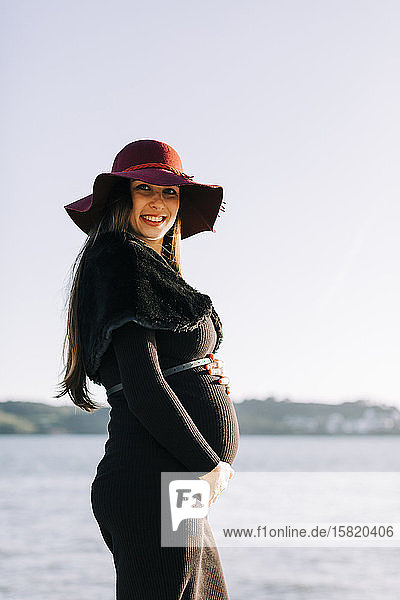 Porträt einer modischen schwangeren jungen Frau am Wasser stehend