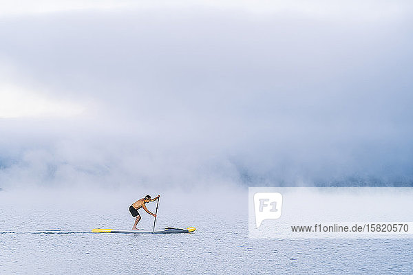 Mann steht beim Paddel-Surfen auf einem See im Nebel auf