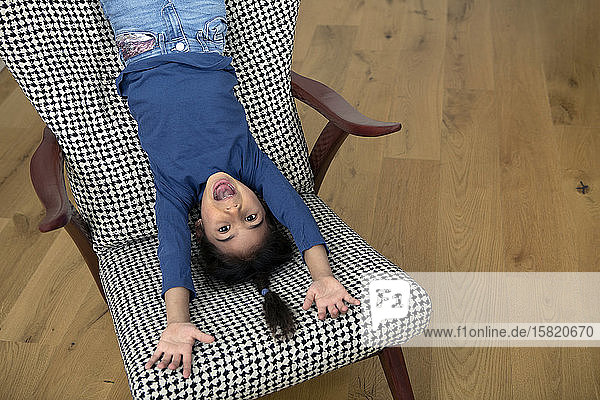 Portrait of little girl hanging upside down from backrest of an armchair having fun
