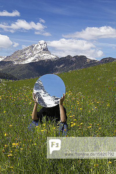 Frau sitzt auf einer Wiese  hinter einem Spiegel versteckt  in dem sich die Berge des Gadertals spiegeln  Südtirol  Italien