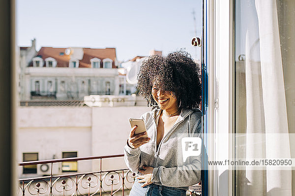 Portrait of happy young woman with curly hair holding cell phone on balcony