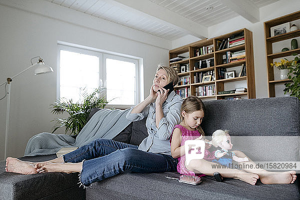 Kleines Mädchen spielt mit Puppe auf dem Sofa  während ihre Mutter telefoniert