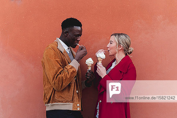 Junges Paar steht an einer Orangenwand und isst Eis