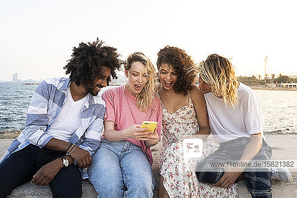 Glückliche Freunde sitzen bei Sonnenuntergang mit Smartphone an der Kaimauer