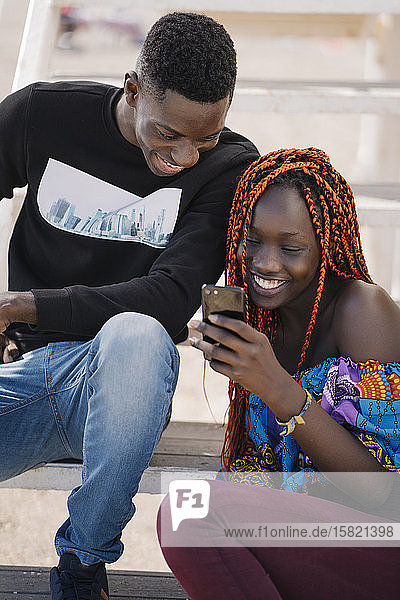 Happy teenage girl and young man sitting on stairs looking at cell phone