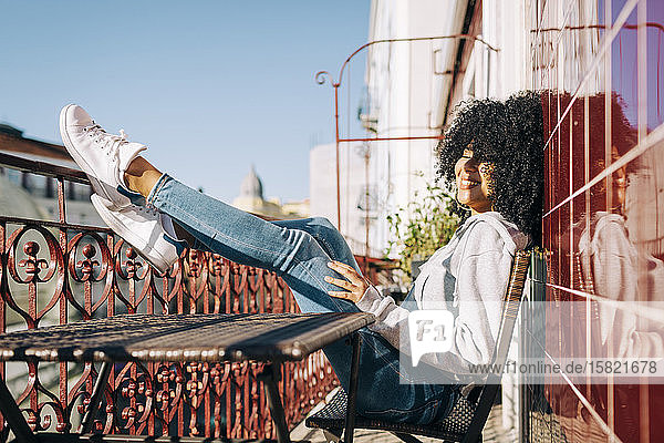 Glückliche junge Frau mit lockigem Haar sitzt auf dem Balkon und genießt den Sonnenschein