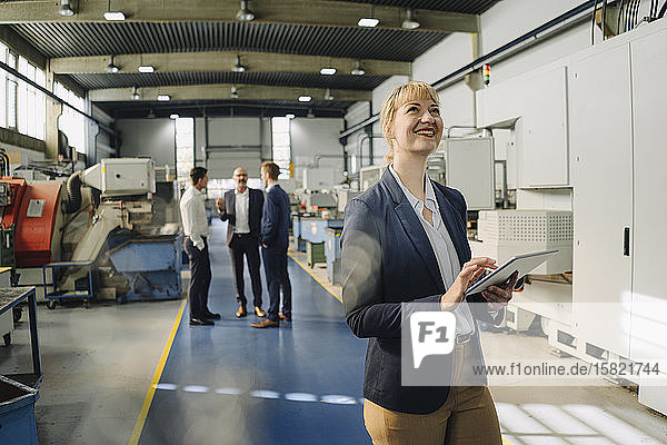 Porträt einer lächelnden Geschäftsfrau mit Tablette in einer Fabrik mit Kollegen im Hintergrund