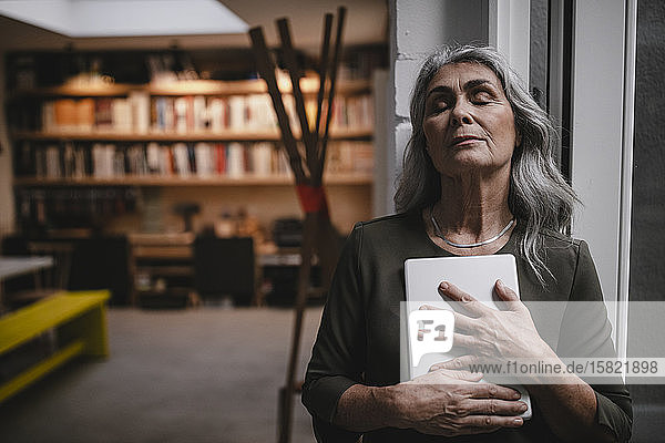 Porträt einer grauhaarigen Geschäftsfrau mit geschlossenen Augen  die in einem Loft-Büro ein Tablett hält