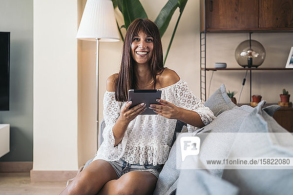 Porträt einer lächelnden jungen Frau  die zu Hause auf der Couch sitzt und Tabletten benutzt