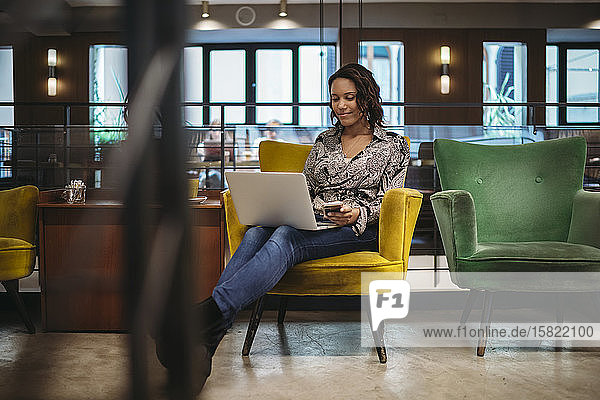 Junge Frau sitzt im Sessel und benutzt Laptop und Smartphone
