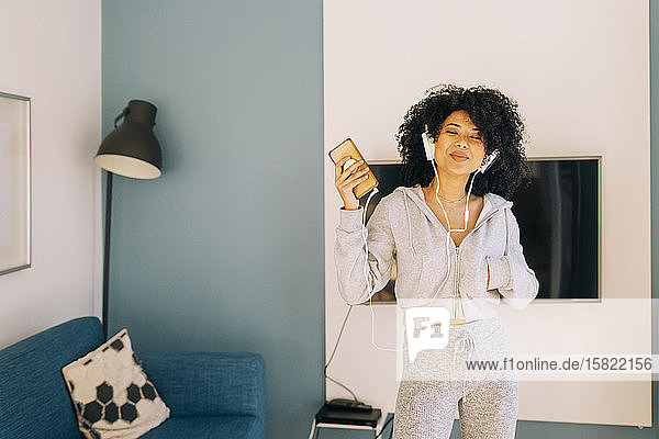 Happy young woman with curly hair listening to music and dancing in living room at home