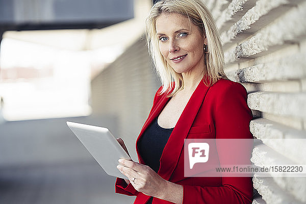 Blonde Geschäftsfrau in rotem Anzug und mit digitalem Tablet in einem Bürogebäude