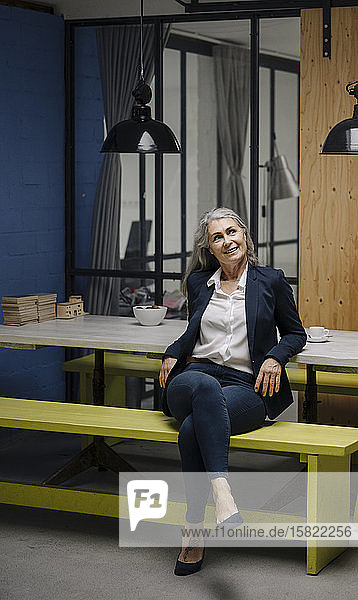 Lächelnde grauhaarige Geschäftsfrau sitzt auf einer Bank in einem Loft-Büro