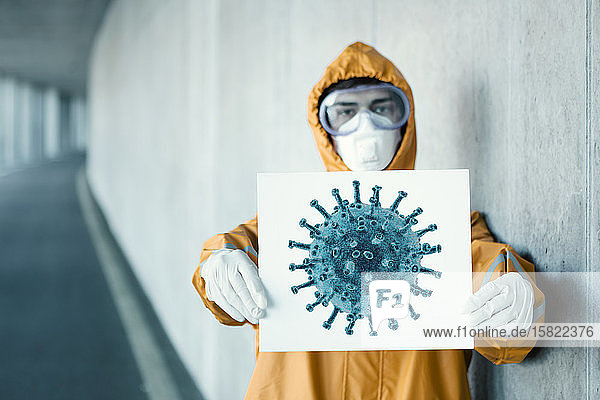 Porträt eines Mannes in Schutzkleidung der ein Schild mit der Darstellung eines Virus hält