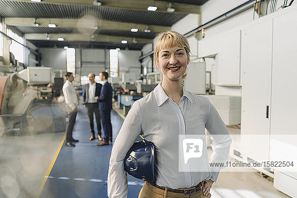 Porträt einer lächelnden Frau mit Schutzhelm in einer Fabrik mit Kollegen im Hintergrund