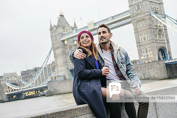 Zwei junge Touristen sitzen mit einem Smartphone an der Wand  mit der London Bridge im Hintergrund