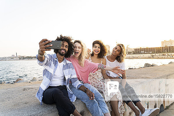 Glückliche Freunde sitzen bei Sonnenuntergang auf der Kaimauer und machen ein Selfie
