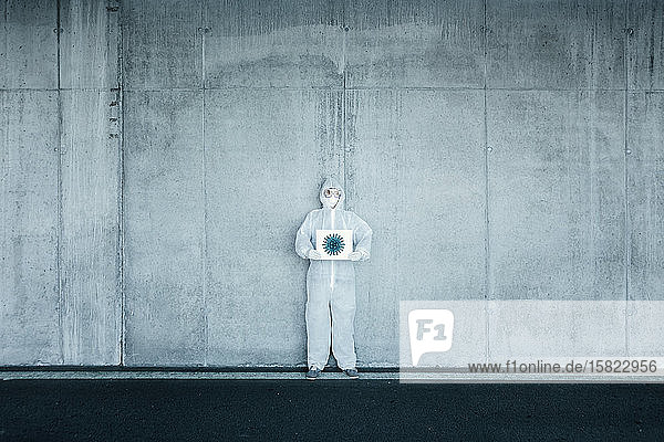 Mann mit Schutzkleidung hält Schild mit der Darstellung eines Virus