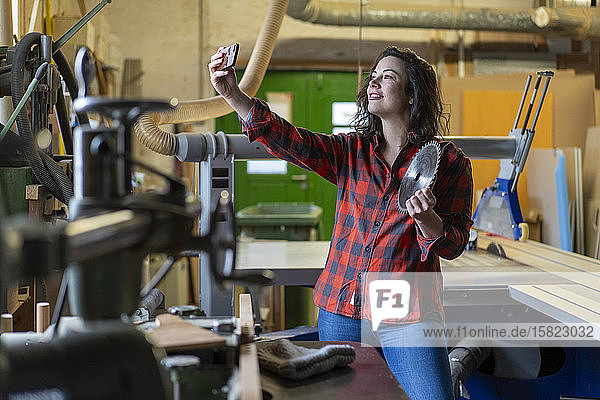 Handwerkerin hält Sägeblatt in der Hand und nimmt in ihrer Werkstatt ein Selfie