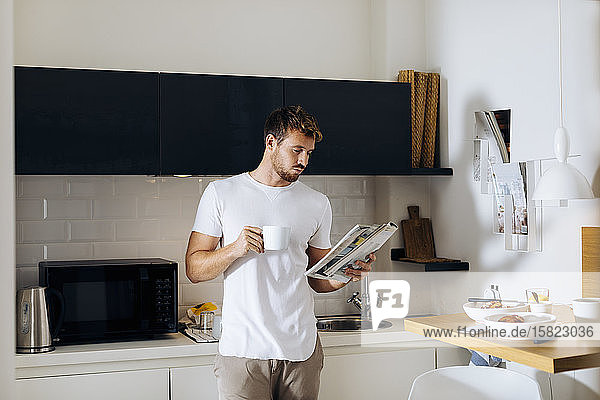 Junger Mann liest eine Zeitschrift und trinkt Kaffee in der Küche