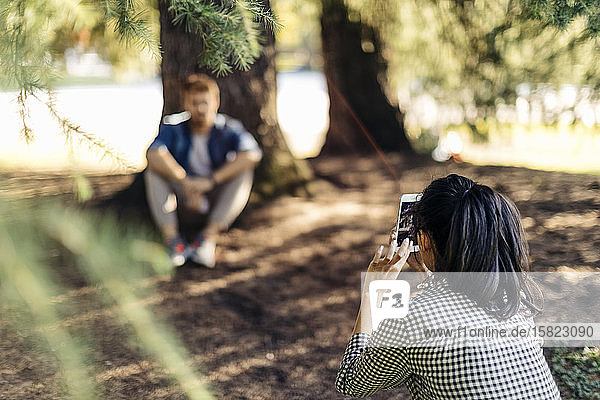 Young woman taking cell phone picture of boyfriend under a tree
