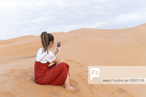 Young woman sitting in sand dune in Sahara Desert using cell phone  Merzouga  Morocco