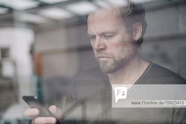Porträt eines Geschäftsmannes  der sein Handy hinter einer Fensterscheibe benutzt