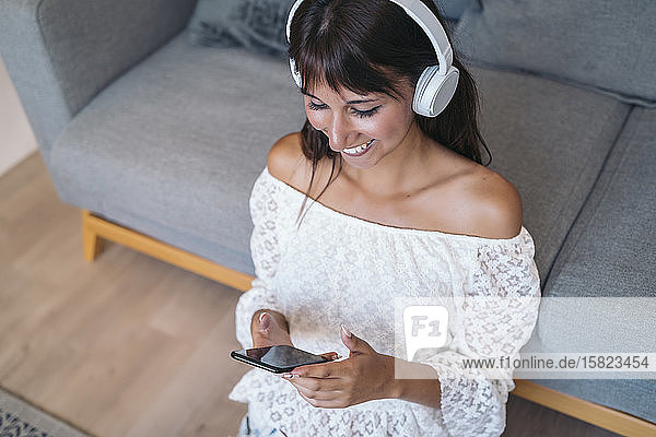 Glückliche junge Frau  die mit Kopfhörern Musik hört und zu Hause im Wohnzimmer auf dem Boden sitzt