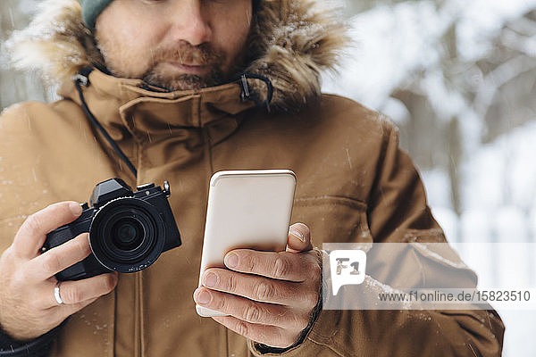 Schnittbild eines Mannes mit Digitalkamera beim Blick auf ein Mobiltelefon im Winter