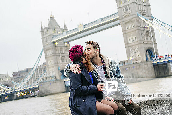 Zwei junge Touristen sitzen mit einem Smartphone an der Wand  mit der London Bridge im Hintergrund