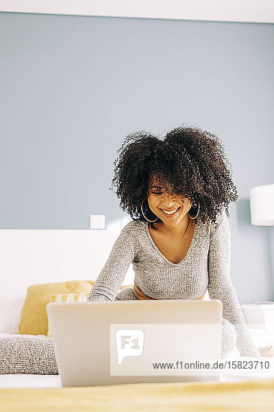 Glückliche junge Frau mit lockigem Haar  die zu Hause einen Laptop auf dem Bett benutzt