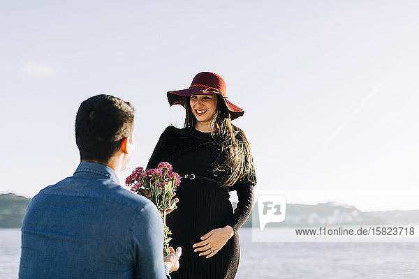 Junger Mann schenkt seiner schwangeren Freundin Blumen an der Uferpromenade