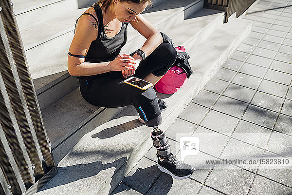 Sportliche junge Frau mit Beinprothese sitzt mit Smartwatch auf einer Treppe in der Stadt