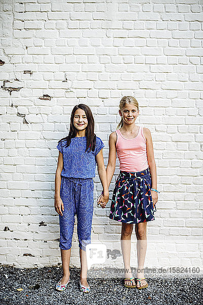 Portrait of two smiling girls standing hand in hand at a wall