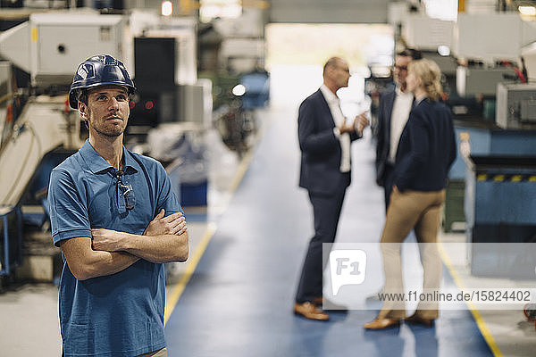 Porträt eines selbstbewussten Arbeiters in einer Fabrik mit Schutzhelm und Geschäftsleuten im Hintergrund